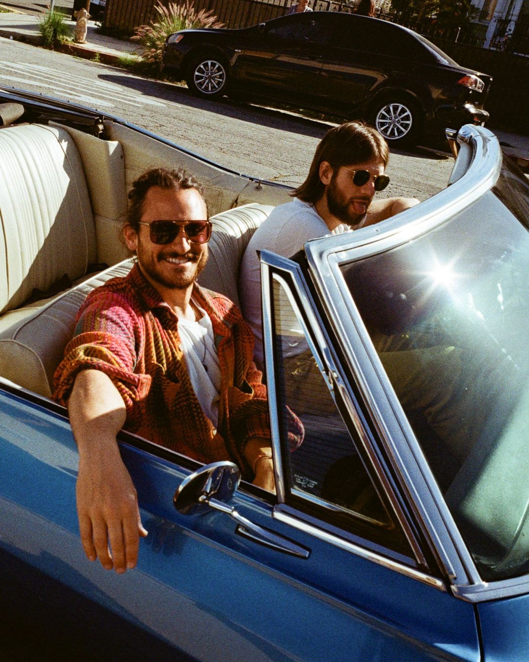 Two adult males ride in a blue convertible car. The passenger is visible with sunglasses on and a dark car is in the background.