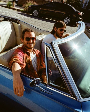 Two adult males ride in a blue convertible car. The passenger is visible with sunglasses on and a dark car is in the background.