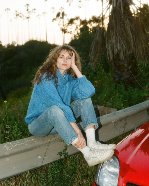 Young woman with bangs sits on a metal guardrail near a red car in a grassy, overgrown area. Trees are silhouetted in the background.