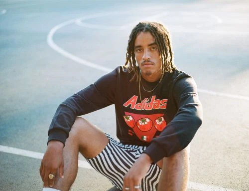 Man in an Adidas sweatshirt and striped shorts sits on a basketball court in the sunlight looking at the camera. Adidas logo visible.
