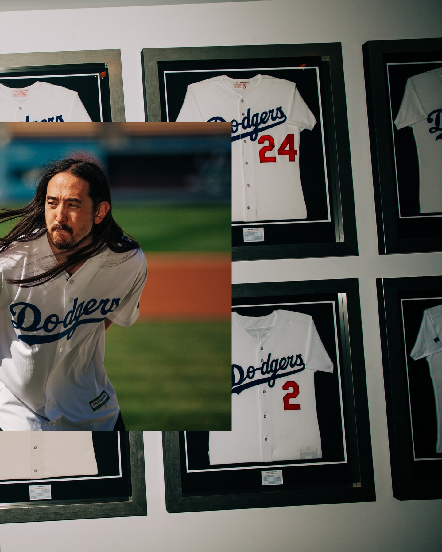 Steve Aoki in a Dodgers baseball uniform with framed Dodgers jerseys on a white wall in the background, creating a sports and music theme.