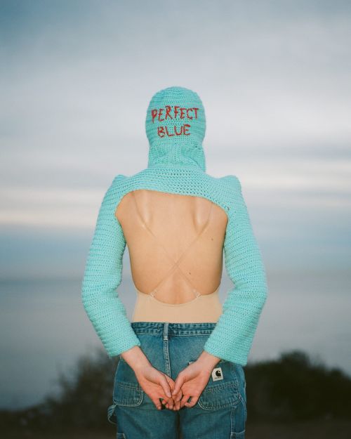 Adult female in jeans and aqua balaclava with arms behind back, facing away, ocean backdrop.