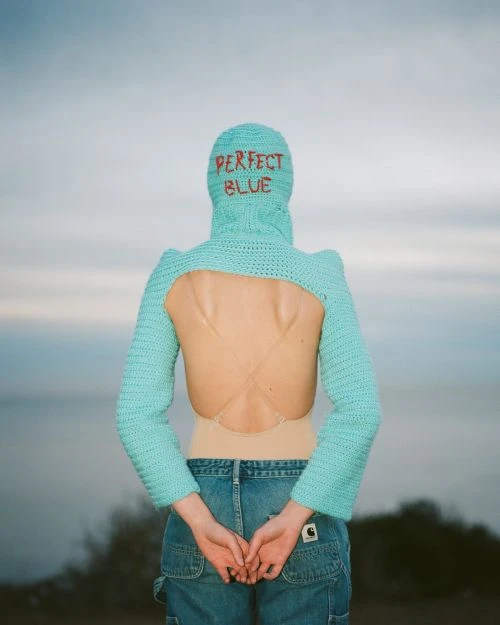 Adult female in jeans and aqua balaclava with arms behind back, facing away, ocean backdrop.