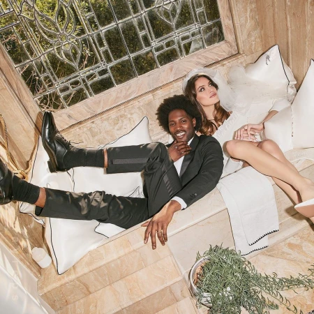 Elegant couple in wedding attire poses on pillows in a luxurious interior setting with natural light from a leaded glass window.