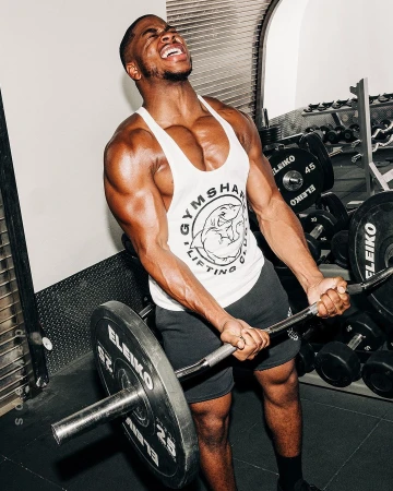 Muscular adult man lifting a barbell with weights in a gym, wearing a Gymshark tank top and black shorts, focusing on his fitness routine.