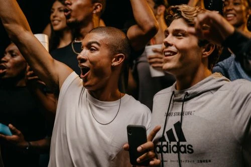 Enthusiastic crowd of diverse young adults cheering at an event, with a man holding a smartphone and wearing an Adidas hoodie visible.