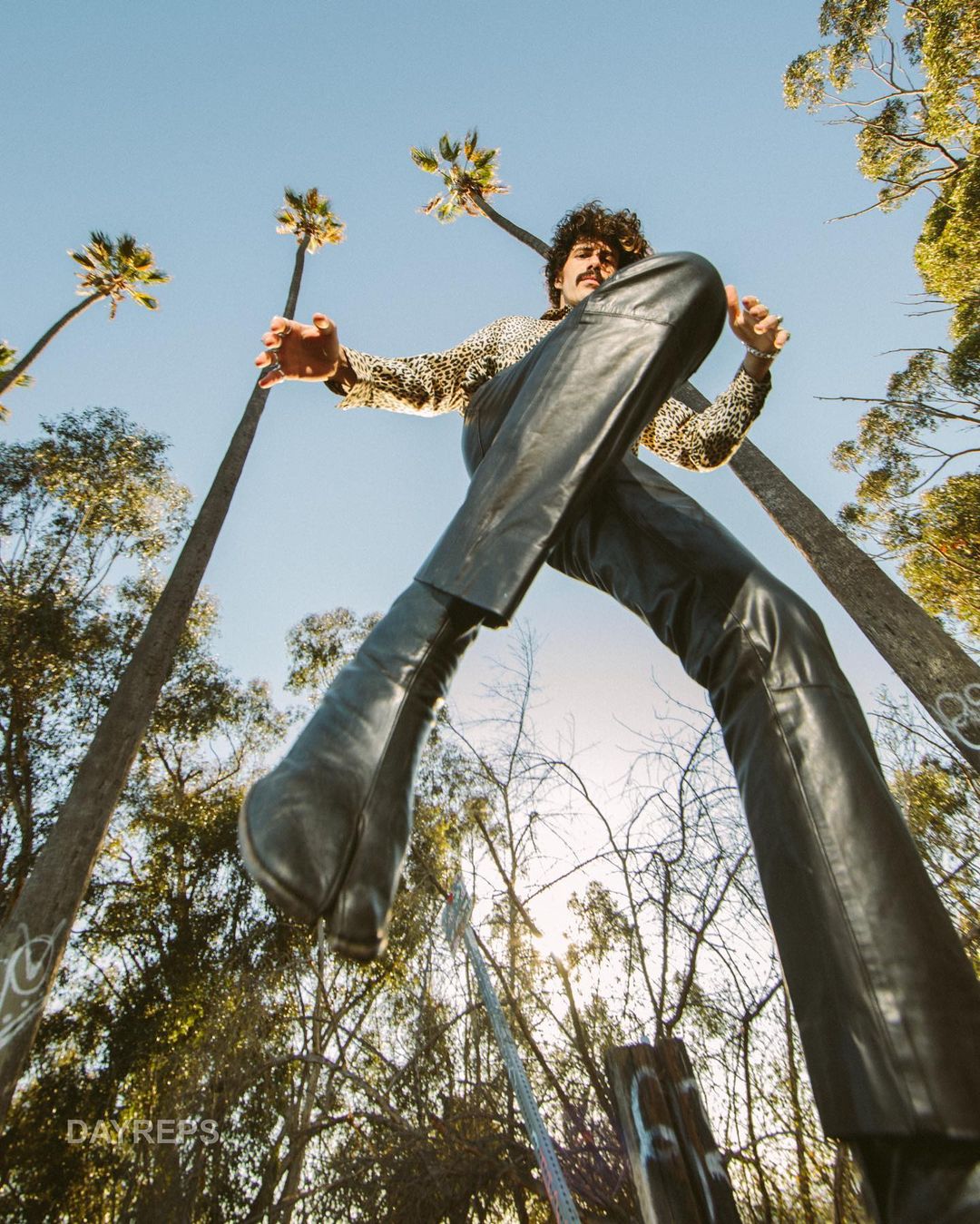 Low-angle view of adult male striding, wearing leather pants, palm trees, and sky in the background in an outdoor setting.