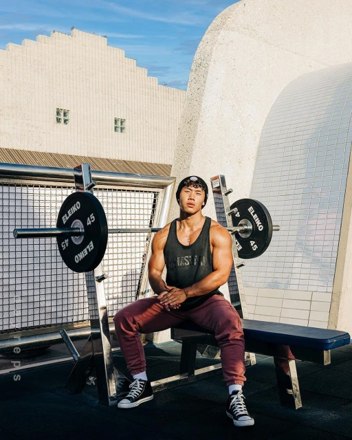 An Asian man in sportswear sits on a weight bench in front of a barbell, set against a backdrop of modern architecture under a blue sky.