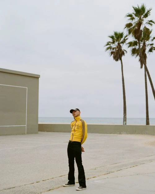 A fashionable adult poses in a yellow track jacket and black jeans on a concrete court with palm trees and ocean in the background.