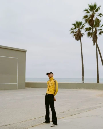 A fashionable adult poses in a yellow track jacket and black jeans on a concrete court with palm trees and ocean in the background.