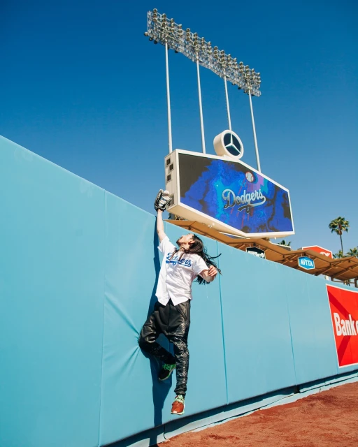 A person in baseball uniform leaps against a stadium wall to catch a ball against the backdrop of stadium lights and a blue sky.