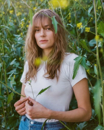 A woman with long wavy brown hair and bangs wearing a white t-shirt, standing amidst tall green foliage and yellow wildflowers.