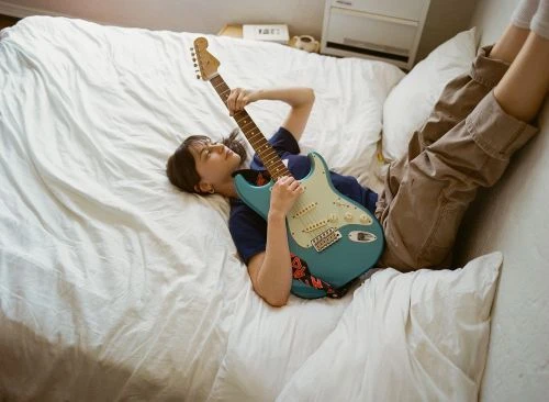 A young woman in a navy shirt and khakis lies on a white bed, playing a light-blue electric guitar in a relaxed, indoor setting.
