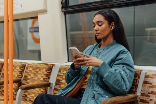 Woman using a mobile phone on a train. She wears a blue bomber jacket and ear buds. The train car has orange patterned seats.