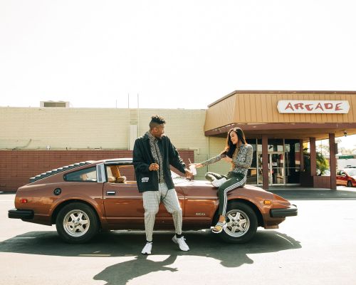 A man and a woman pose on a brown sports car parked outside an arcade. The image is bathed in bright daylight.