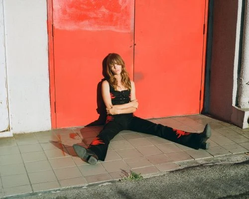 A young woman with long brown hair sits in front of a bright red door wearing a black outfit with red details and black boots.