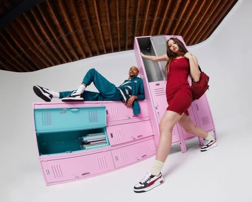 Two models strike poses in front of pink and teal lockers in a light, airy studio. A woman in a red dress and a man in a teal tracksuit wear Puma sneakers.