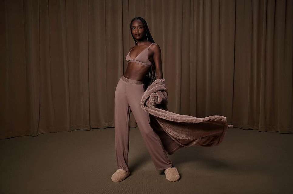 Adult woman poses in mauve UGG sleepwear, featuring a bralette, pants, robe, and slippers against a neutral curtain backdrop, fashion advertising.