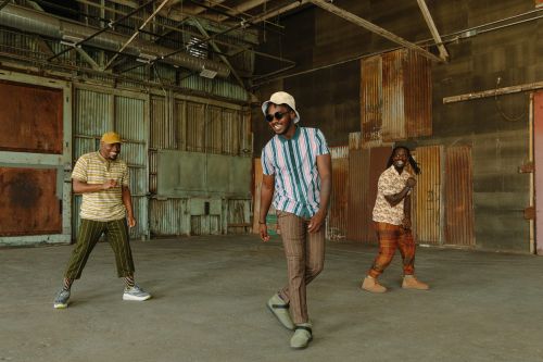 Three African American men in patterned shirts and pants dance in an open warehouse with natural light and a high ceiling.