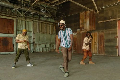 Three African American men in patterned shirts and pants dance in an open warehouse with natural light and a high ceiling.