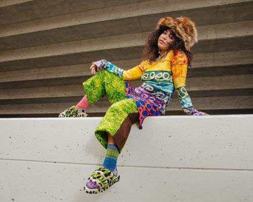 A person with curly hair wears a multi-color top, green pants, and furry hat and shoes while sitting on a concrete wall outdoors.