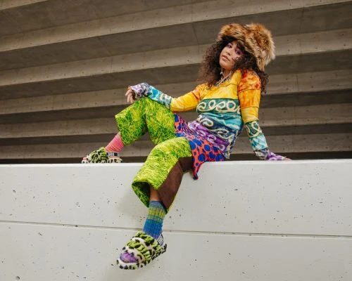 A person with curly hair wears a multi-color top, green pants, and furry hat and shoes while sitting on a concrete wall outdoors.
