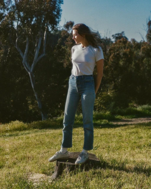 Woman in t-shirt and jeans stands on tree stump in field. Forest and blue sky background on sunny day. Fashion editorial outdoors.