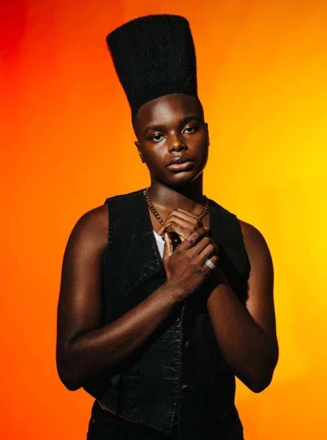 Portrait of a stylish Black man with a flattop haircut, wearing a vest and necklace, standing against an orange-toned studio backdrop.