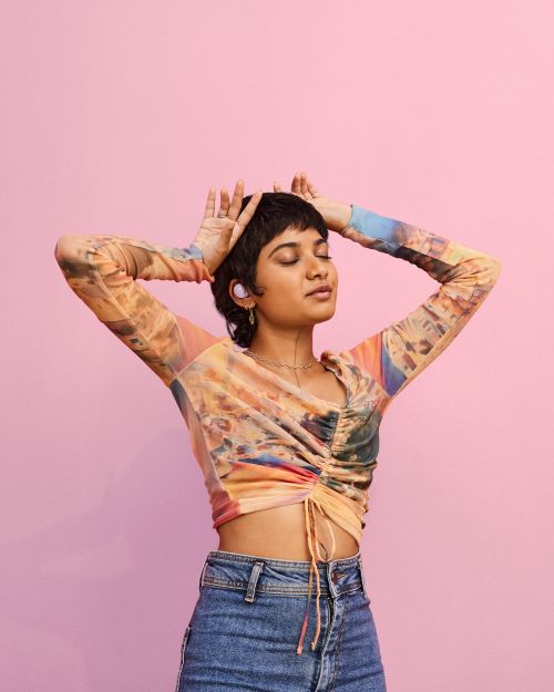 Young woman with short hair poses with her hands above her head and eyes closed in front of a pink background in a studio shot.