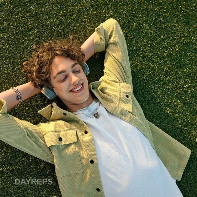 Young adult male relaxes on green grass wearing headphones, light green jacket, and white t-shirt, smiling with eyes closed in the sunlight.