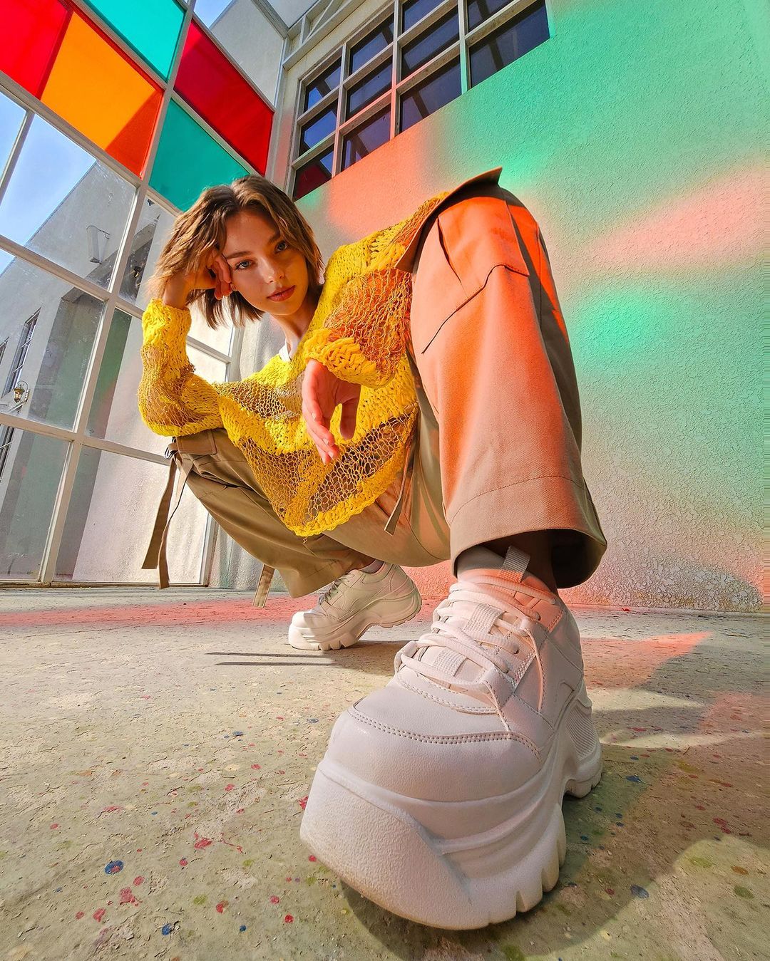 Eye-level shot of a young adult with short hair and wearing a yellow shirt, khaki pants, and white platform sneakers in urban setting.