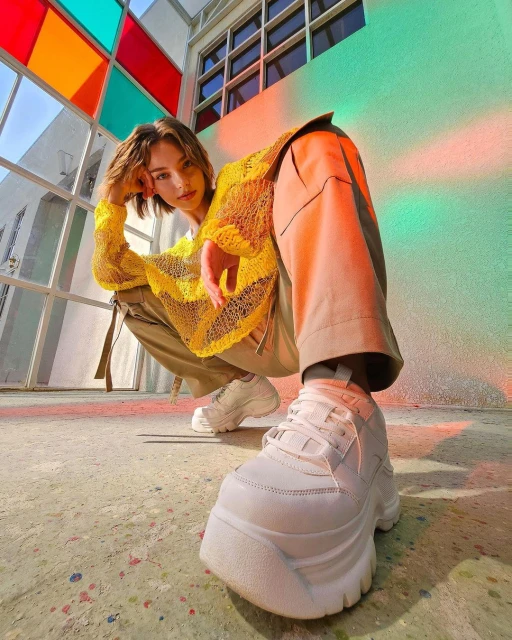 Eye-level shot of a young adult with short hair and wearing a yellow shirt, khaki pants, and white platform sneakers in urban setting.