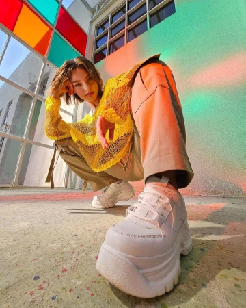 Eye-level shot of a young adult with short hair and wearing a yellow shirt, khaki pants, and white platform sneakers in urban setting.