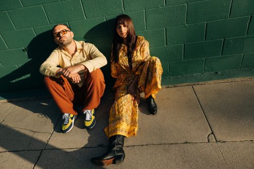 A stylish couple poses against a green brick wall in sunlight. The woman wears a yellow print dress and boots; the man is in orange pants.