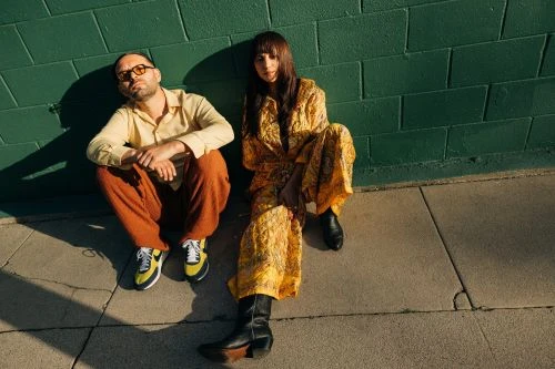 A stylish couple poses against a green brick wall in sunlight. The woman wears a yellow print dress and boots; the man is in orange pants.