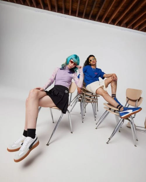 Two young adults in athletic wear pose in a photo studio sitting on chairs, with a white backdrop and wooden ceiling beams.