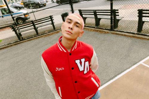 Young adult with buzzcut poses in red and white varsity jacket in urban park setting with benches and parked cars in the background.