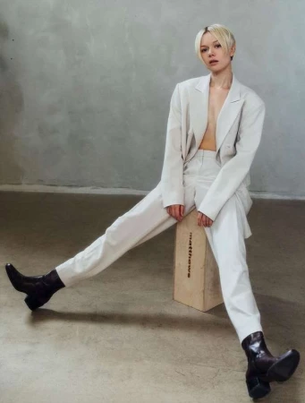 Blonde woman wearing a cream pantsuit and dark boots seated on a wooden apple box against a concrete wall