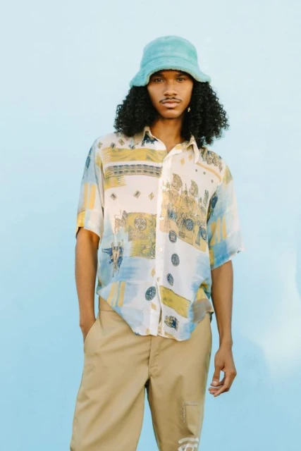 Young adult male in short-sleeved shirt and bucket hat poses against a light blue background. The subject is centered in the frame.