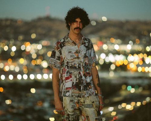 Man with curly dark hair and mustache wearing a newspaper-print short-sleeve shirt against a blurred city bokeh background