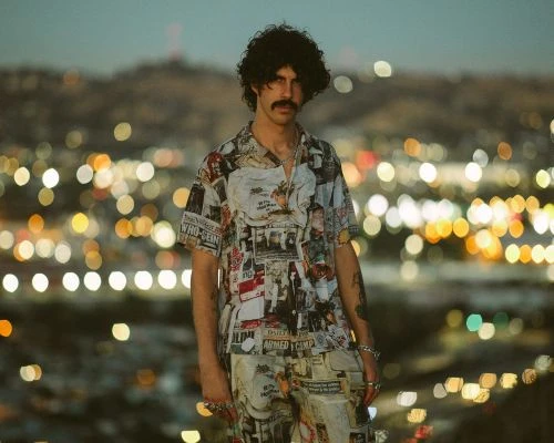Man with curly dark hair and mustache wearing a newspaper-print short-sleeve shirt against a blurred city bokeh background