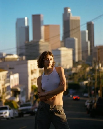 Young woman with short brown hair wearing a white tank top and dark pants posing outdoors with a city skyline background.
