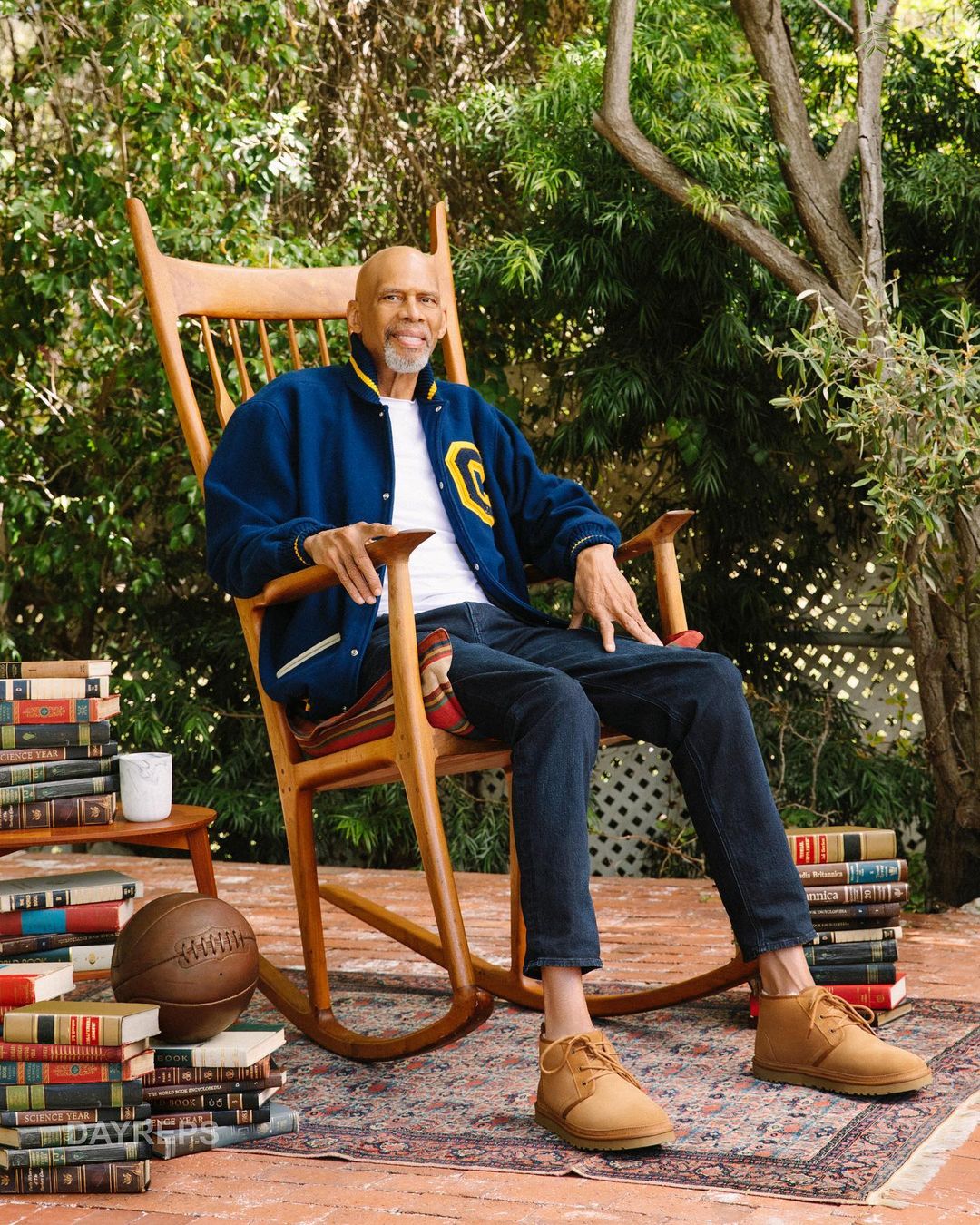 Adult male sitting in a wooden rocking chair on a patterned rug surrounded by stacks of books outside in a garden setting.
