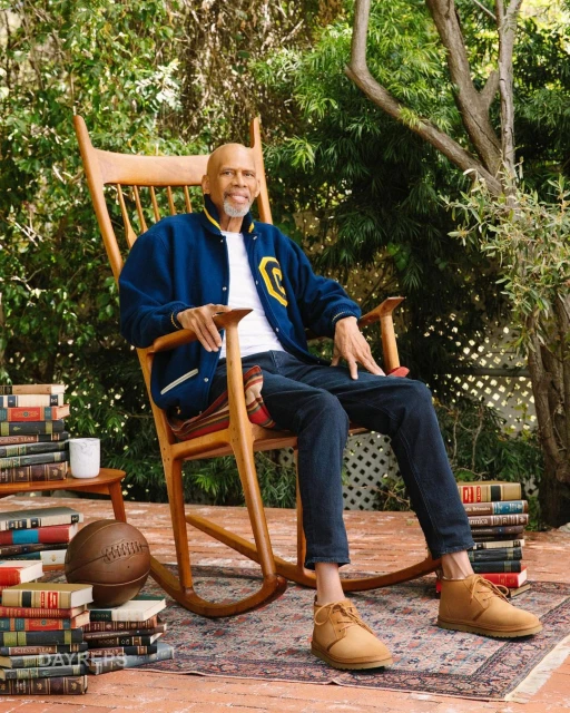 Adult male sitting in a wooden rocking chair on a patterned rug surrounded by stacks of books outside in a garden setting.
