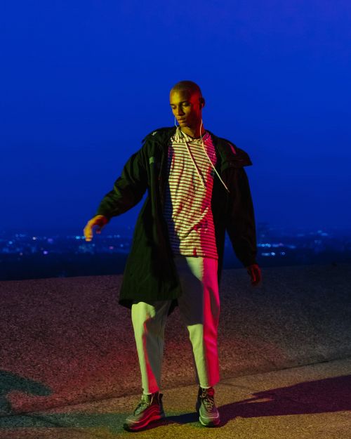 Young adult in trendy streetwear stands outdoors at night with city lights in background under red and blue lighting. 