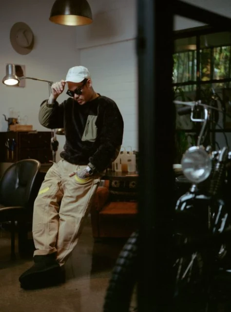 Adult male wearing sunglasses and a cap, resting his arm on a counter with a motorbike in the background, and an indoor setting.
