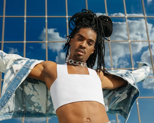 Stylish Black male model poses in a white crop top and bleached denim jacket, with a blue sky and grid-patterned window background.