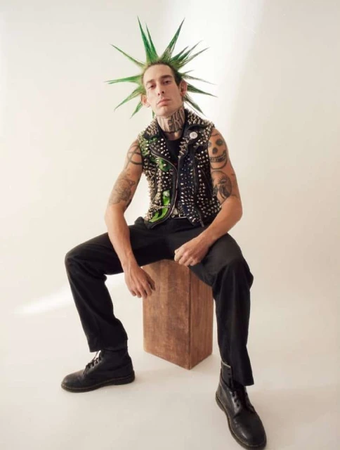 Adult male with green spiked hair sits on a wooden box wearing a studded vest, black pants, and boots against a light background.