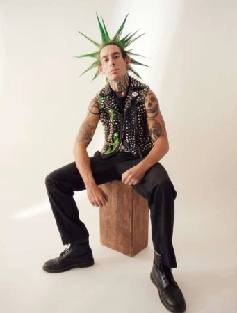 Adult male with green spiked hair sits on a wooden box wearing a studded vest, black pants, and boots against a light background.