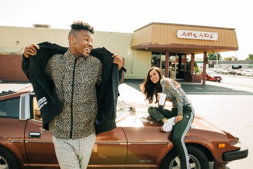 Two adults on a brown car parked outside of an arcade building on a sunny day, one man opening his jacket, one woman sitting on the car.